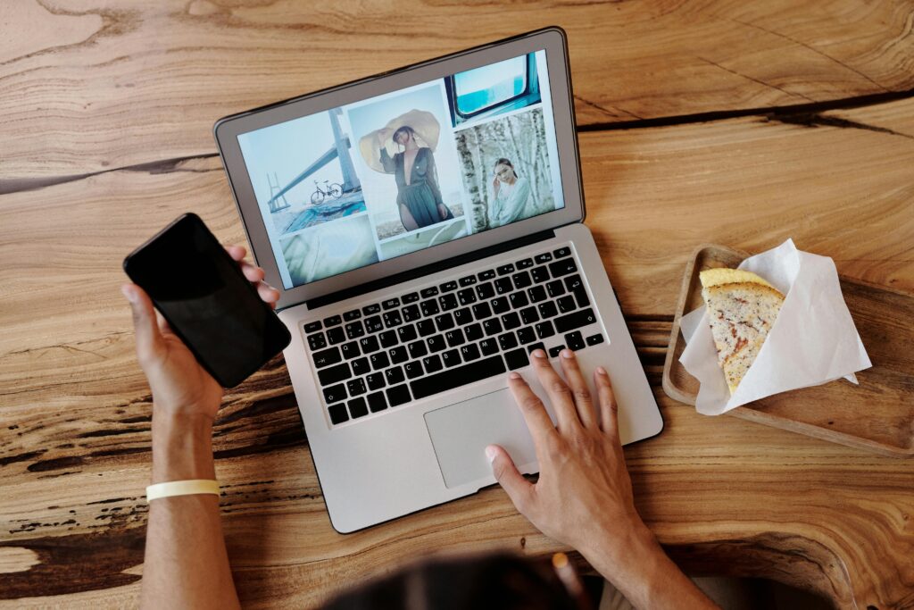 Person uses smartphone and laptop while enjoying a sandwich at a wooden table, showcasing multitasking and technology use.